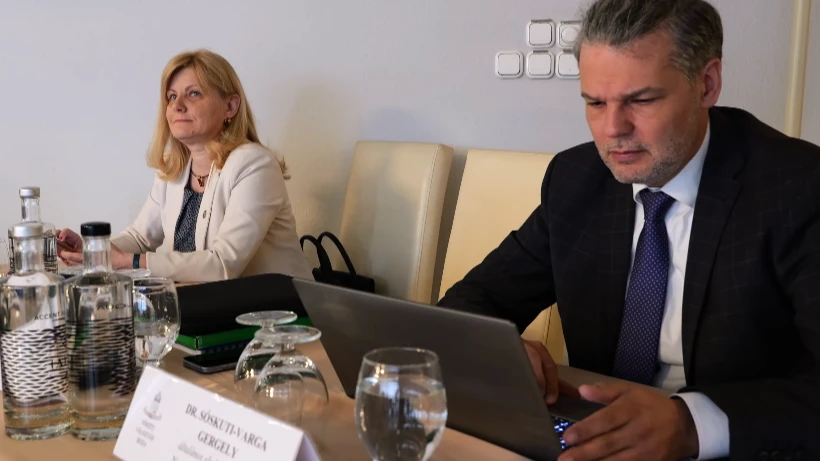 Két hivatalosan öltözött személy ül egy tárgyalóasztalnál. A férfi laptopon dolgozik, előtte névtábla és vizespoharak láthatók, mellette egy nő figyelmesen ül. A környezet formális, megbeszélésre utal.