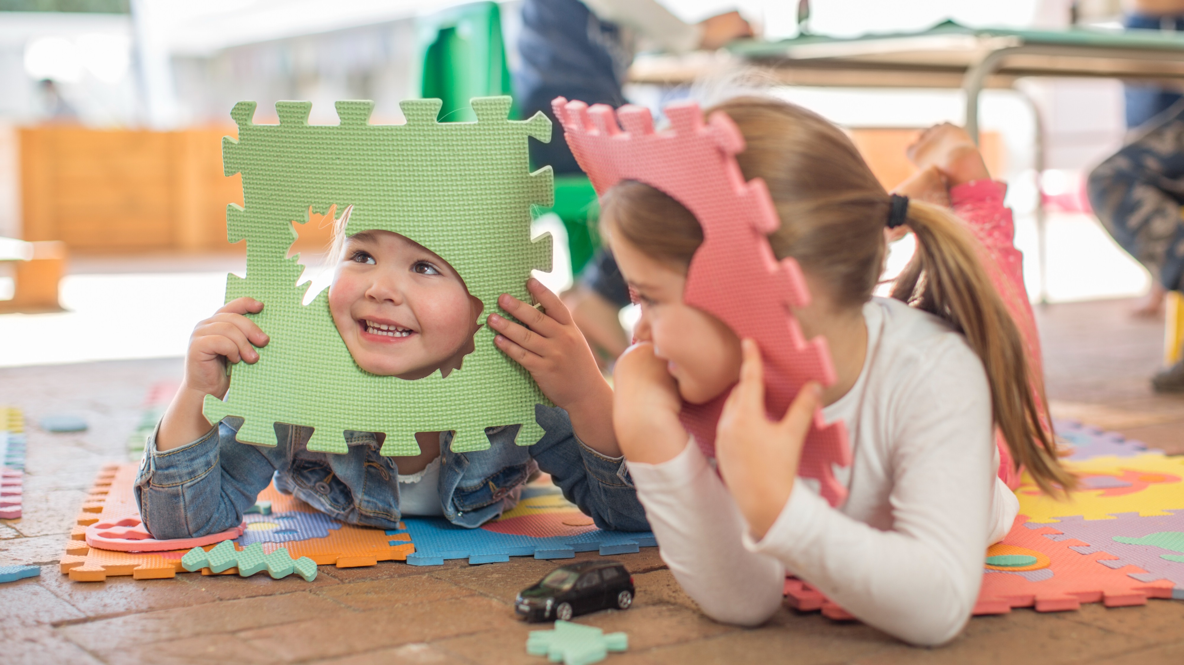 A fotón két kisgyermek fekszik hason egy színes, habszivacs kirakós szőnyegen egy világos óvodai vagy játszószobai környezetben. A háttérben további gyerekek játszanak asztaloknál.