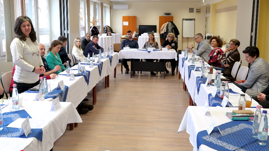 Egy világos tárgyalóteremben U alakban elrendezett asztalok körül több ember ül megbeszélésen; egy nő állva beszél a csoporthoz, az asztalokon víz, jegyzetek és névtáblák láthatók.