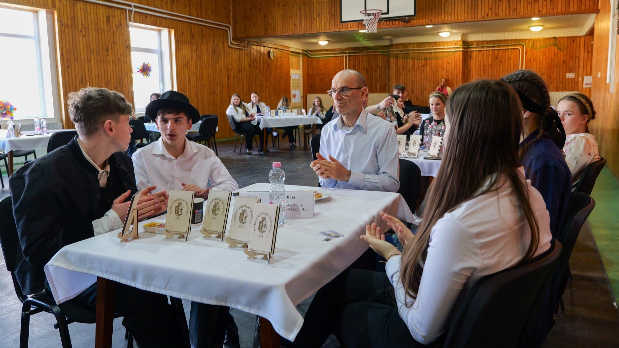 Egy fa burkolatú tornateremben fiatalok ülnek fehér terítővel letakart asztaloknál. Az egyik asztalnál diákok és egy idősebb férfi beszélgetnek, előttük kis táblák és névtáblák láthatók, a háttérben pedig más csapatok figyelnek.