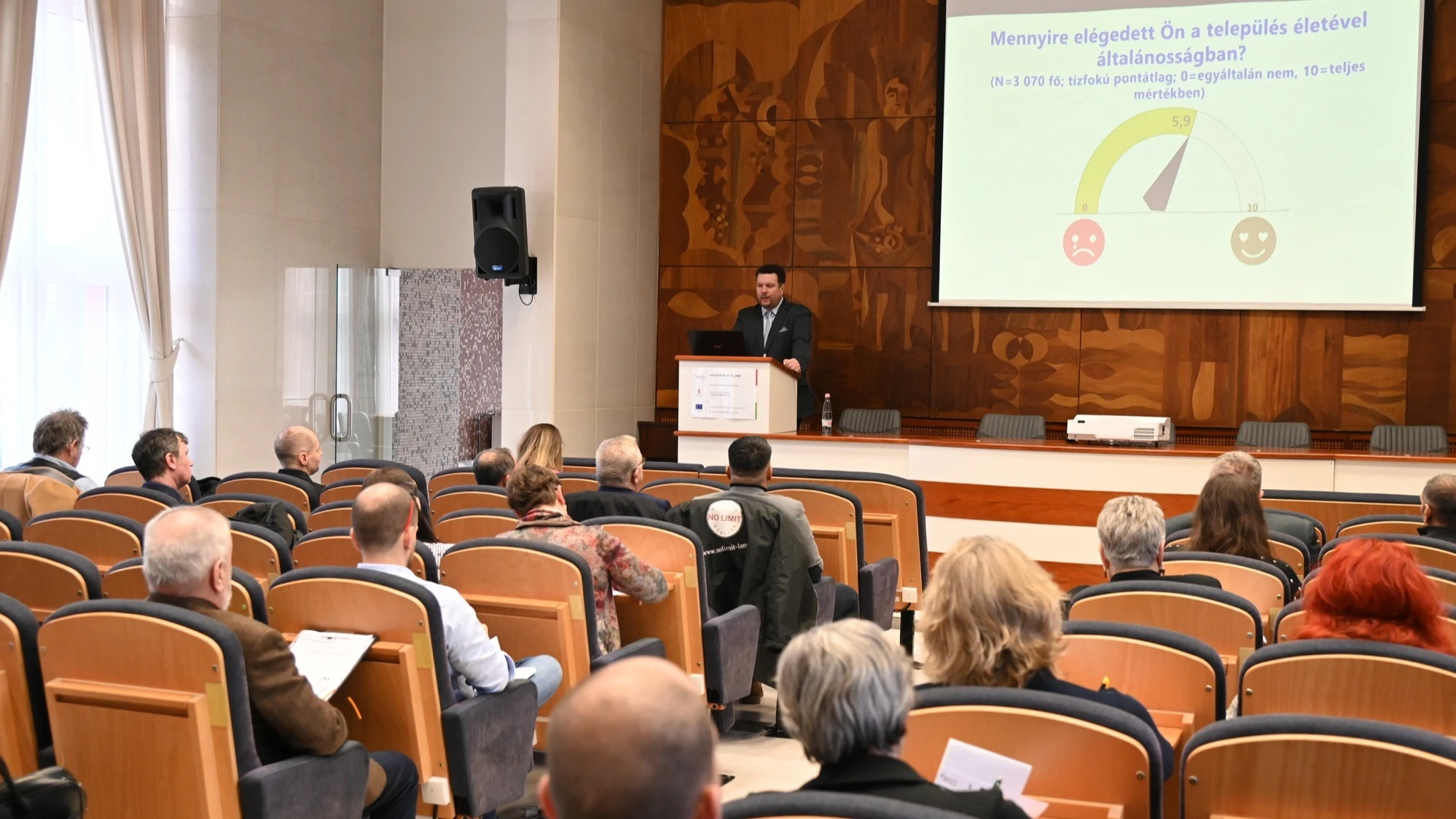 Egy előadótermet látunk, hátulról fotózva. Sorokban világos fa székek vannak, bennük kb. 20–30 fős hallgatóság ül.  Elöl egy öltönyös férfi áll a pódium mögött, és előadást tart laptopról. 