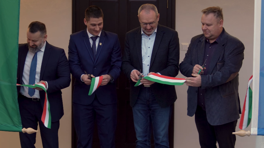 Négy öltönyös férfi áll egymás mellett egy bejárat előtt, és nemzeti színű szalagot vágnak át ollóval. Az esemény ünnepélyes szalagátvágásra utal, háttérben ajtó és zászlók láthatók.