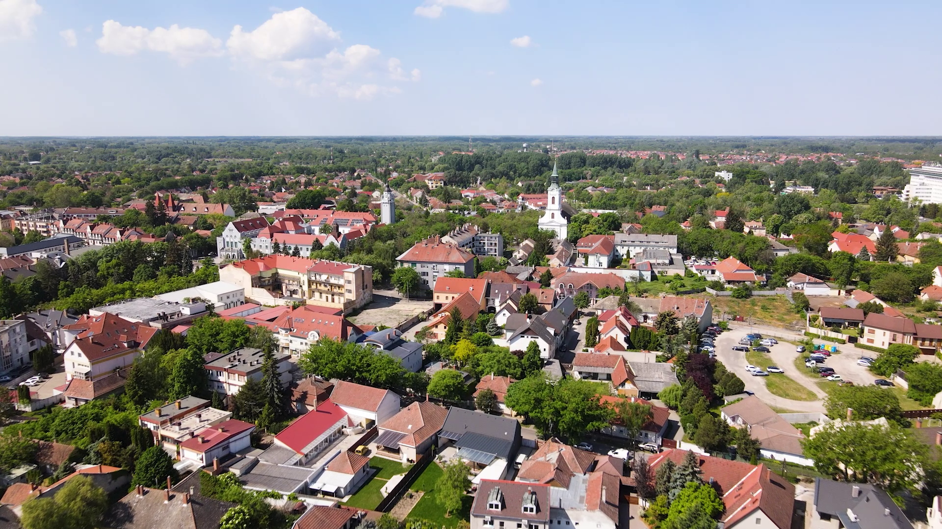 A fotón egy alföldi kisváros madártávlati képe látható: piros cseréptetős családi házak, zöld fákkal sűrűn borított utcák és kisebb középületek terülnek el a távolba. A kép középső részén egy fehér templomtorony emelkedik ki a házak közül.