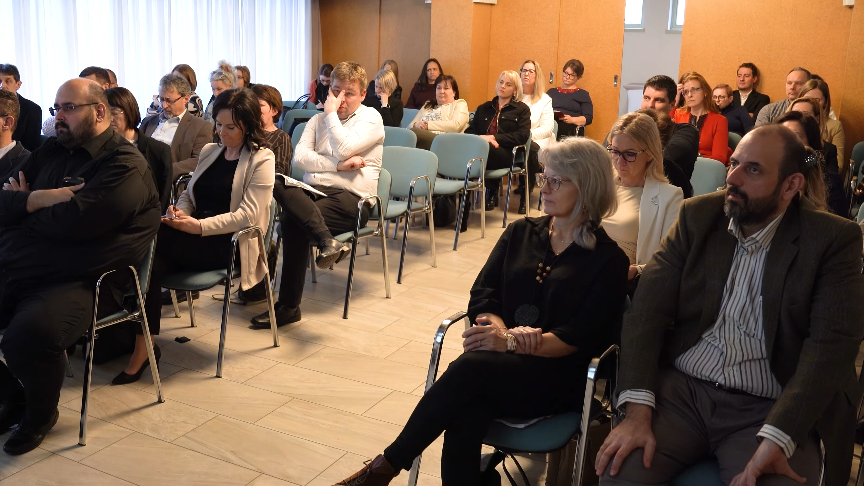 Egy konferenciateremben több tucat ember ül székeken, egymás felé fordulva. A résztvevők többsége jegyzetel vagy figyelmesen hallgatja az előadót, aki a képen nem látható. A hangulat formális, szakmai rendezvényre utal.