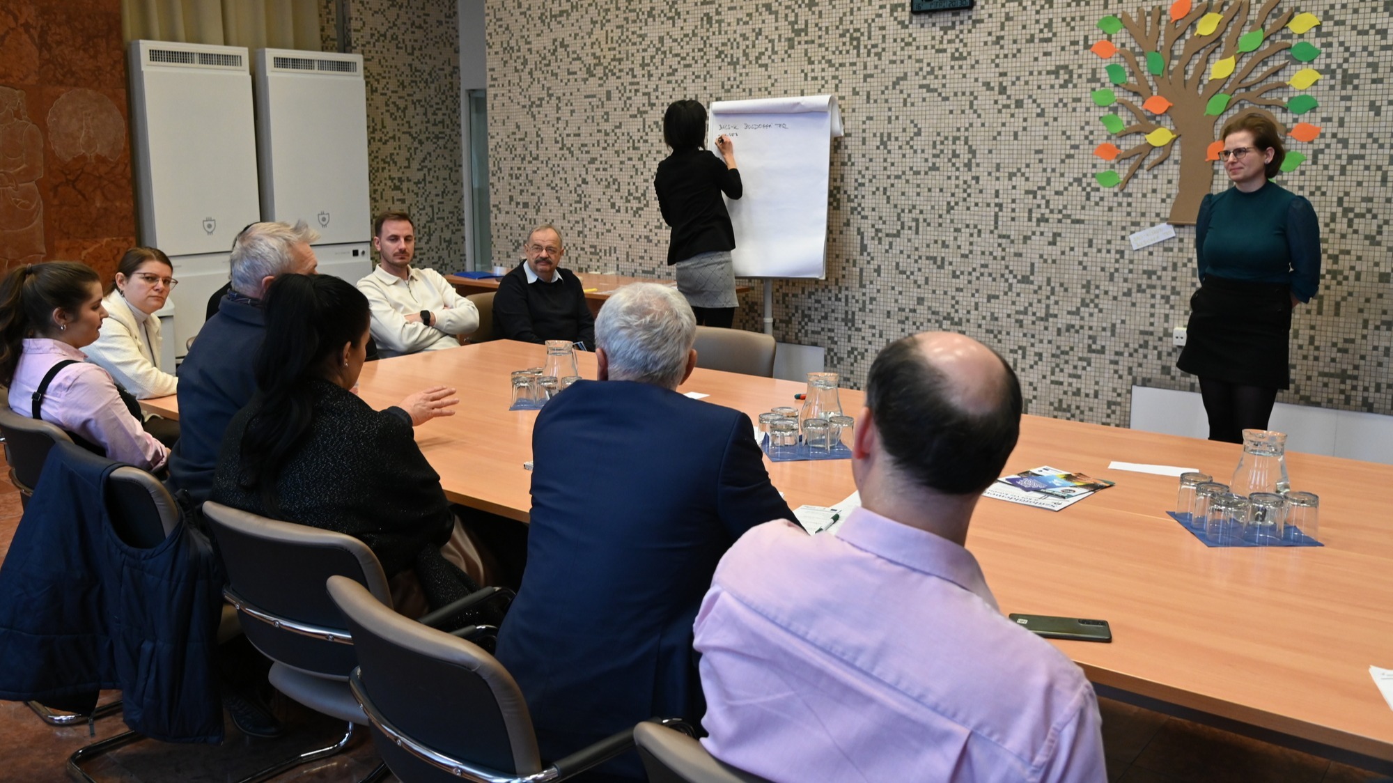 Egy tárgyalótermet látunk, hosszú asztallal és körülötte ülő, körülbelül 10–12 fős csoporttal.  A résztvevők egymás felé fordulva beszélgetnek, egy kiscsoportos megbeszélés zajlik. 