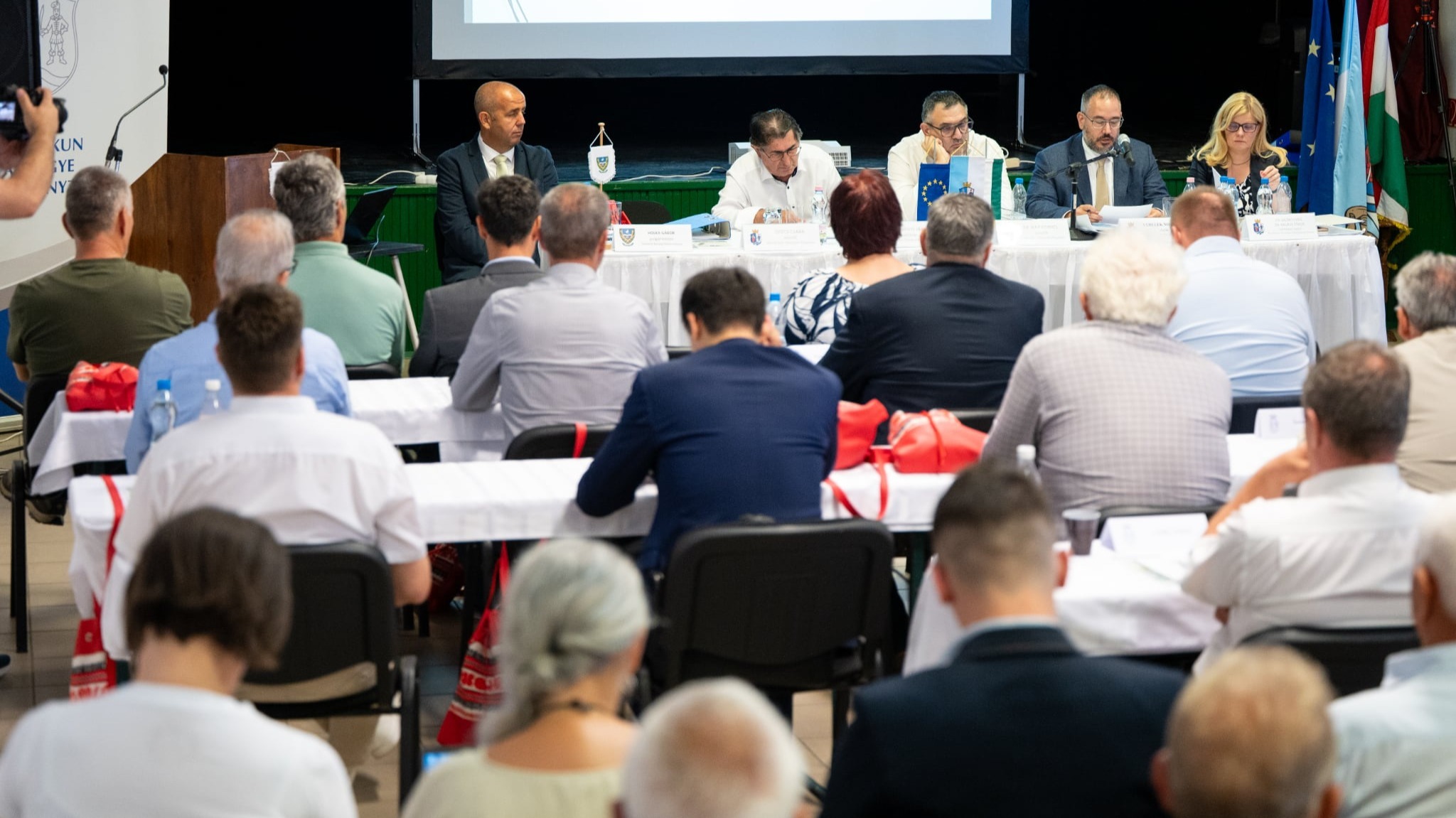 A képen a közgyűlés tagjai láthatók asztalaiknál, hátulról fotózva.