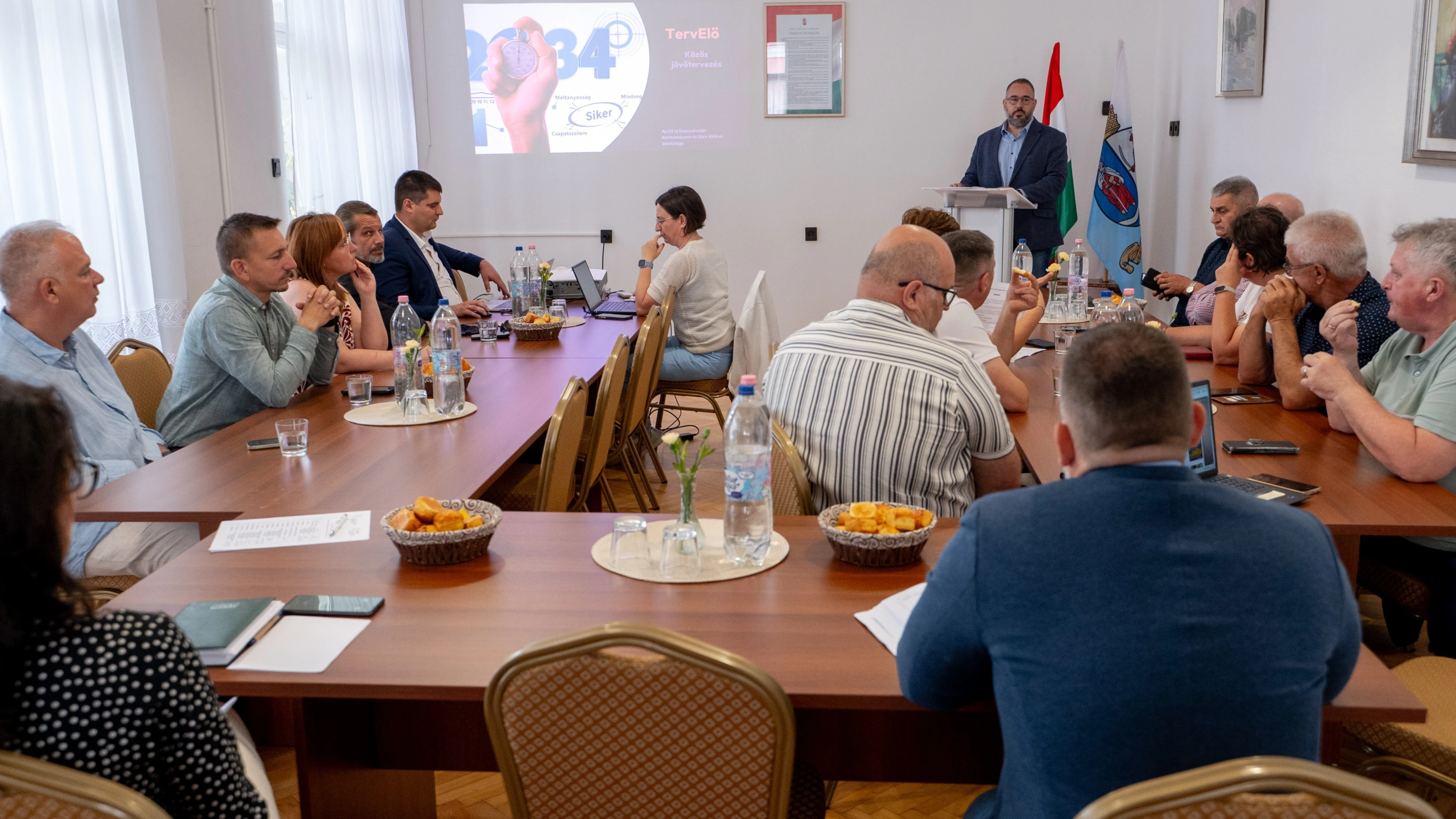 Emberek ülnek asztalok körül egy teremben, ge yvalaki előadást tart, a többiek őt figyelik.