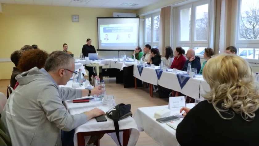 Egy teremben egymással szembe fordított asztaloknál ülnek az emberek, egy hölgy áll, és beszél.