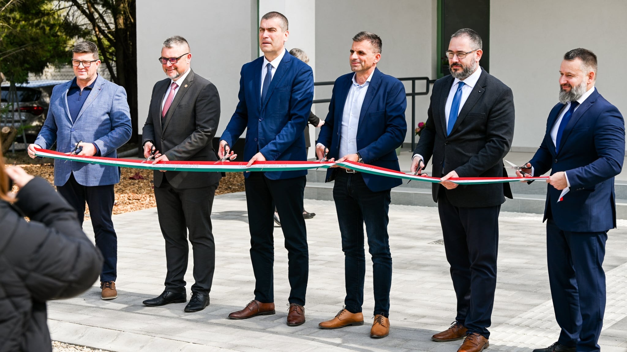 A képen 6 úriember látható, akik ünnepélyesen átvágják a nemzeti színű szalagot.