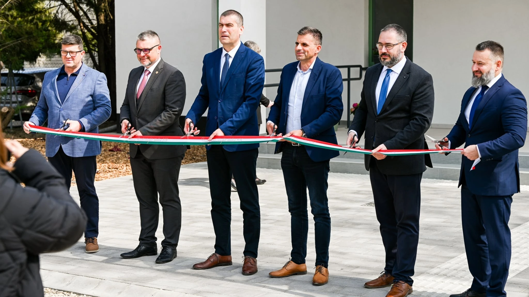 A képen 6 úriember látható, akik ünnepélyesen átvágják a nemzeti színű szalagot.