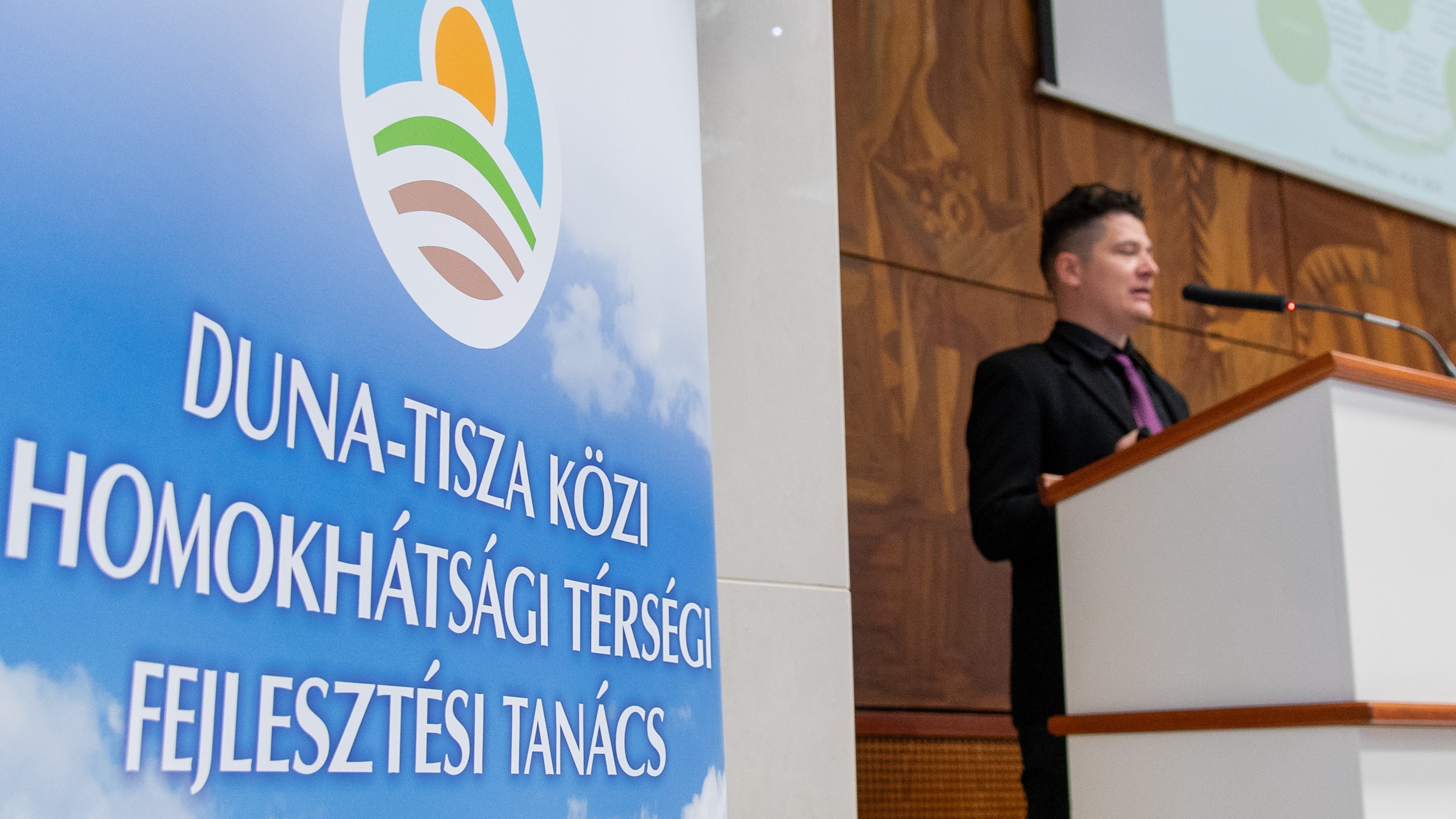 A kép bal oldalán egy roll up a Duna-Tisza Közi Homokhátsági Térségi Fejlesztési Tanács felirattal. A Kép jobb oldalán egy férfi előadó a pulpitusnál.