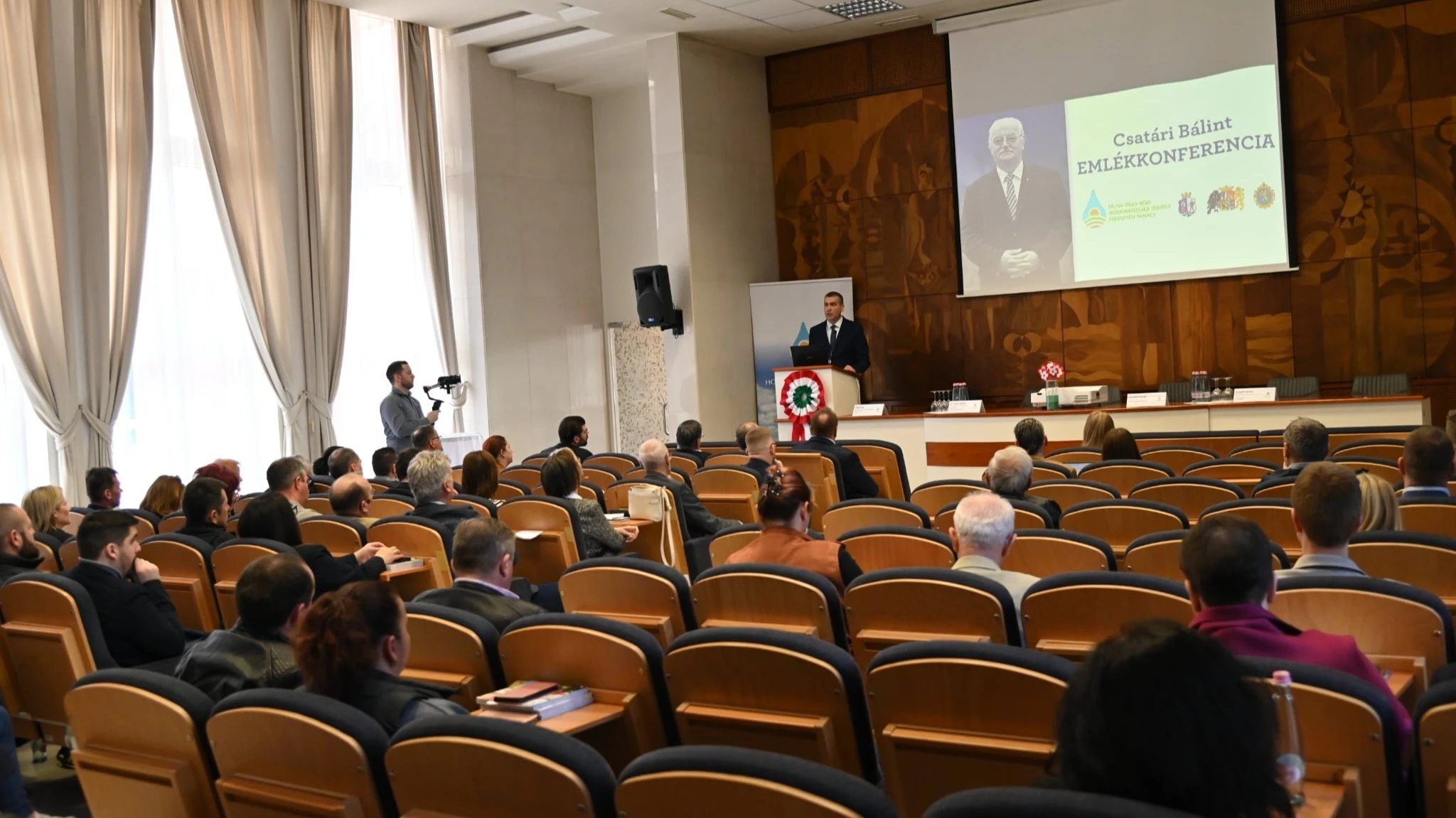 A képen rendezvényterem, ahol a jelenlévők az éppen beszélő férfire figyelnek.