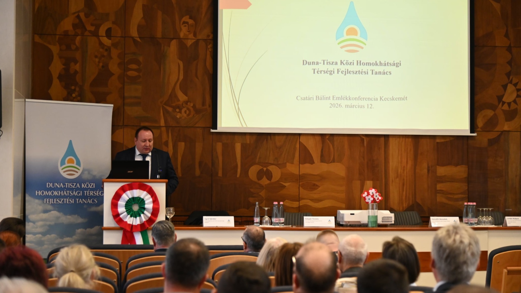 A teremben lévő emberek hallgatják a konferenciaterem élén álló úr beszédét.