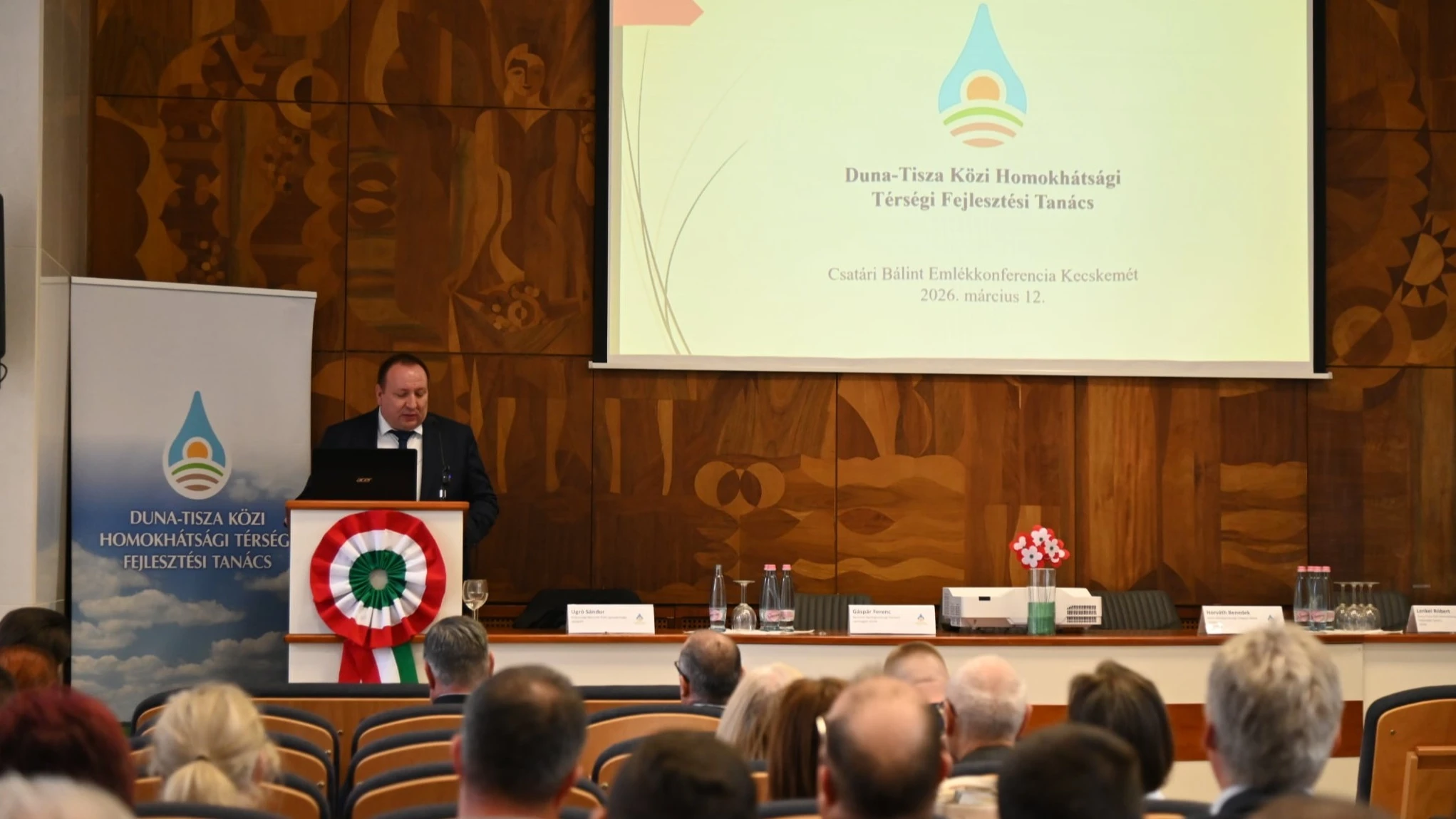 A teremben lévő emberek hallgatják a konferenciaterem élén álló úr beszédét.