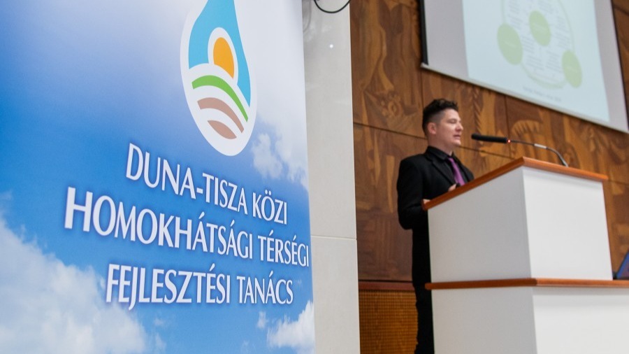 A képen a Duna-Tisza Közi Homokhátsági Térségi Fejlesztési tanács logója látszik az előtérben, mellett egy férfi előadást tart. 