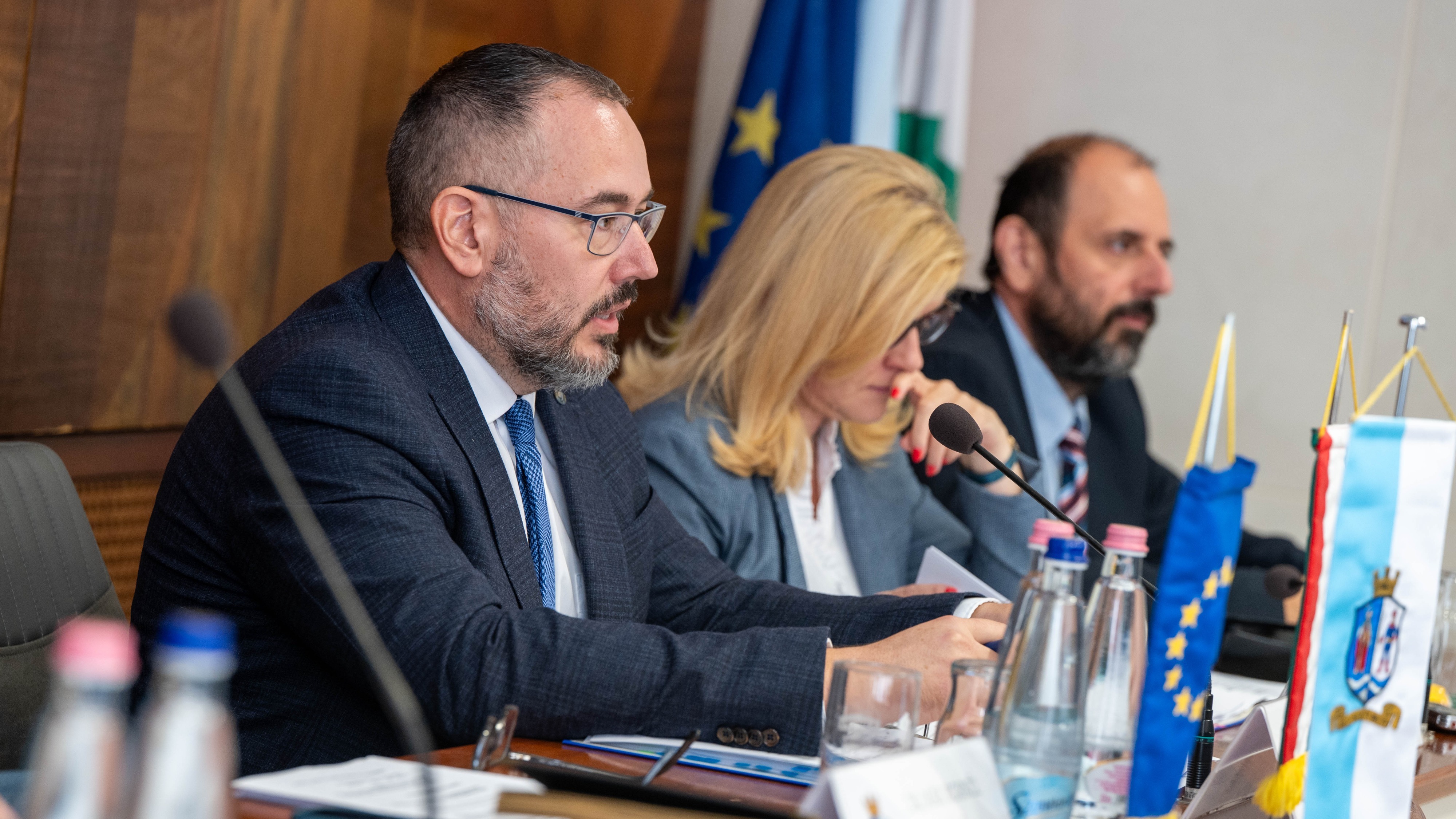 Egy pódiumon ül három személy, előtérben egy férfi aki beszél, mellette egy hölgy és egy másfik férfi.